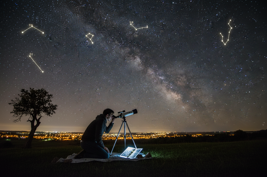Beginner stargazer using small telescope with star chart under night sky filled with visible constellations